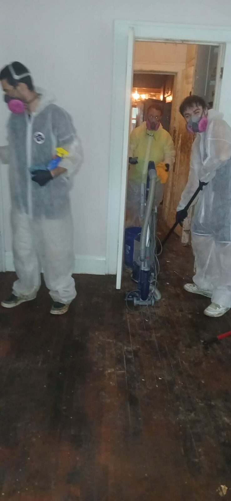 Three crew members in protective suits and pink respirators cleaning a stained wood floor